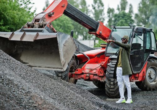 Stylish african american man in hat and sunglasses posed outdoor in rain against tractor with a bucket.